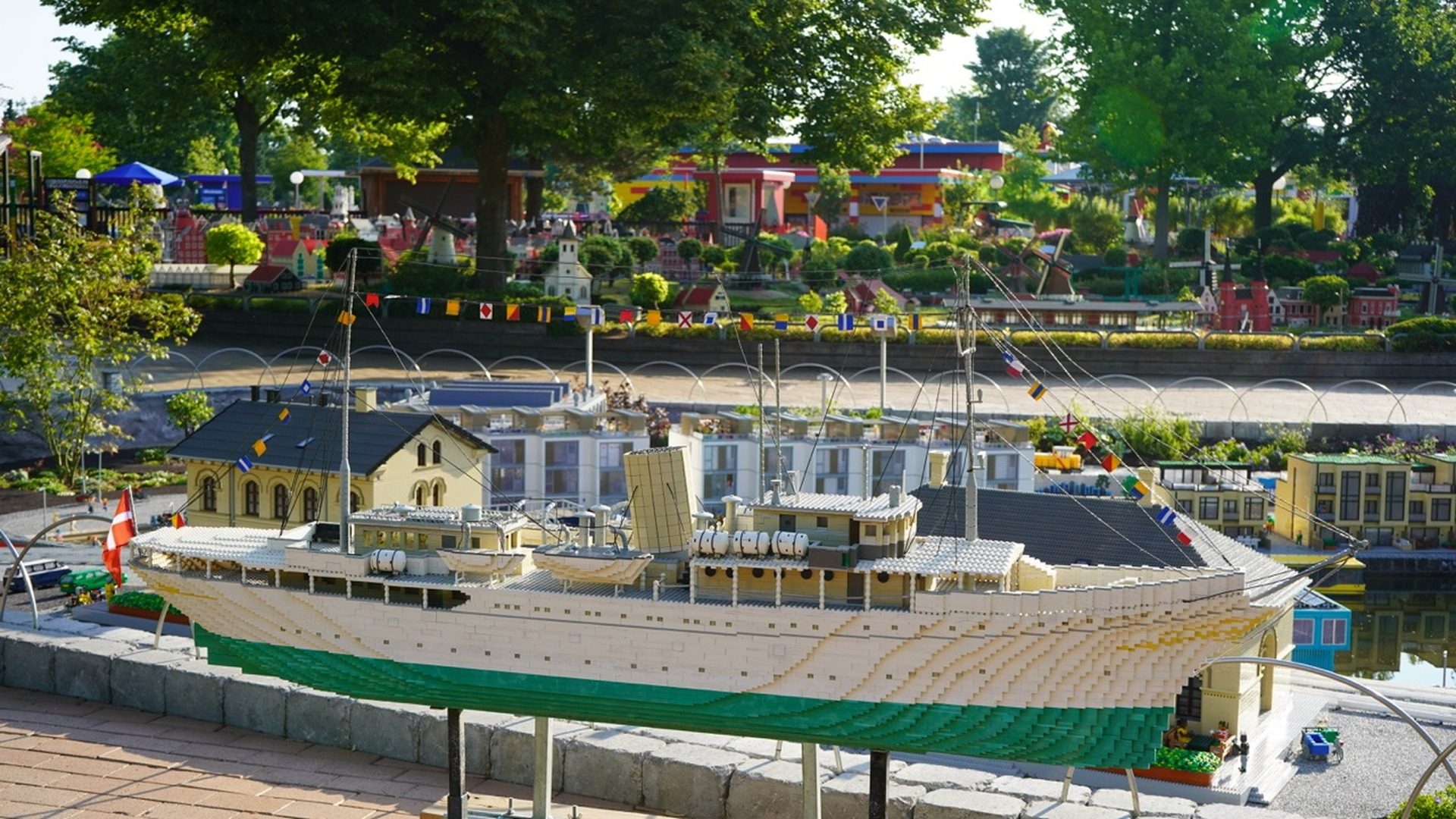 Large LEGO ship model with cream and green hull docked at LEGOLAND harbor with miniature buildings and landscaping.