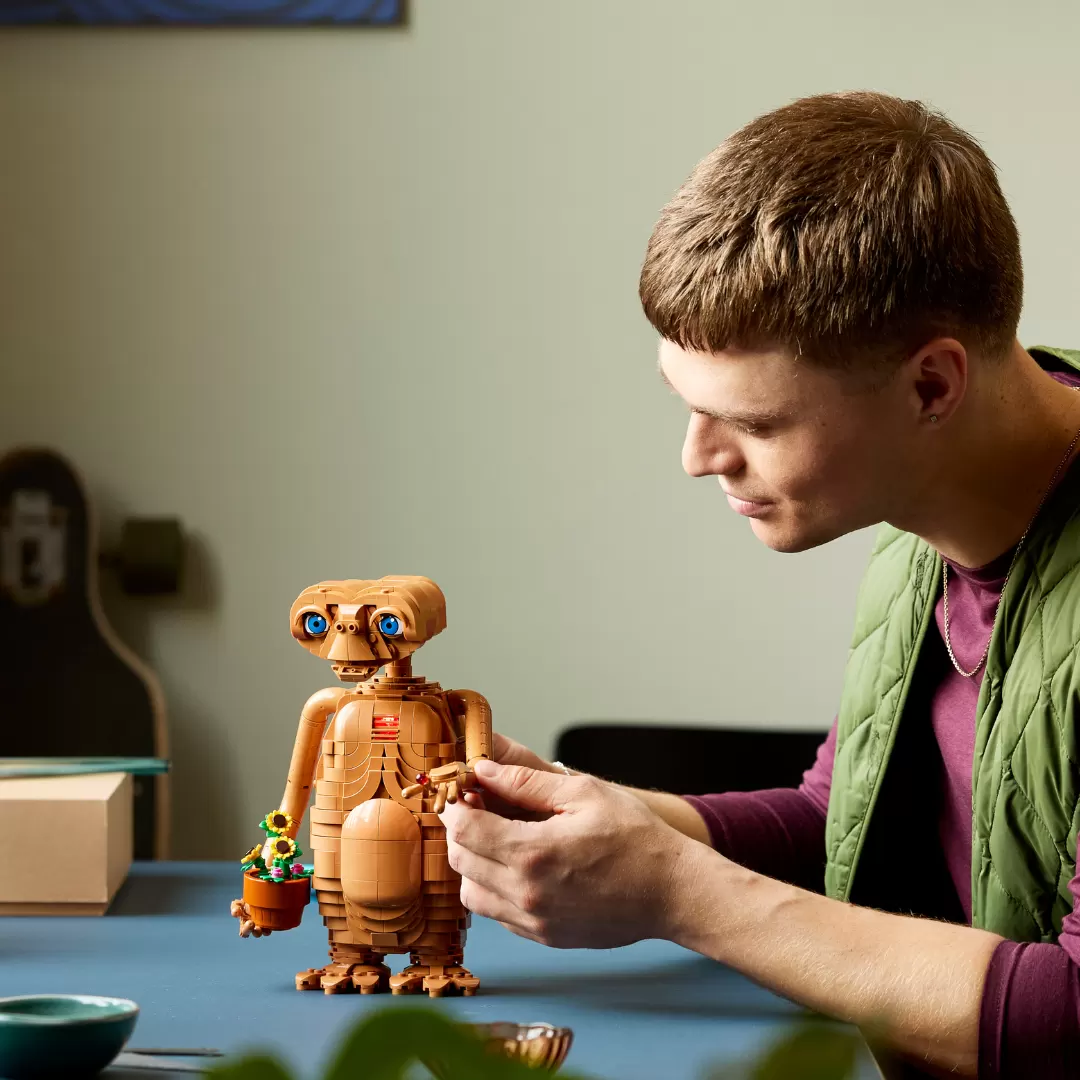 A young man examines a tan LEGO E.T. figure with glowing blue eyes and a small colorful creature beside it on a blue table.