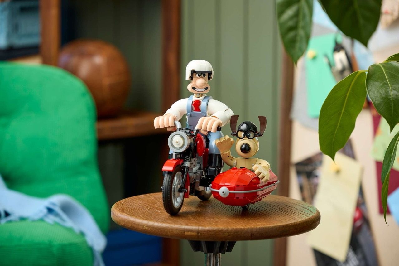 LEGO Wallace minifigure on red motorcycle with sidecar containing tan dog minifigure on wooden table.