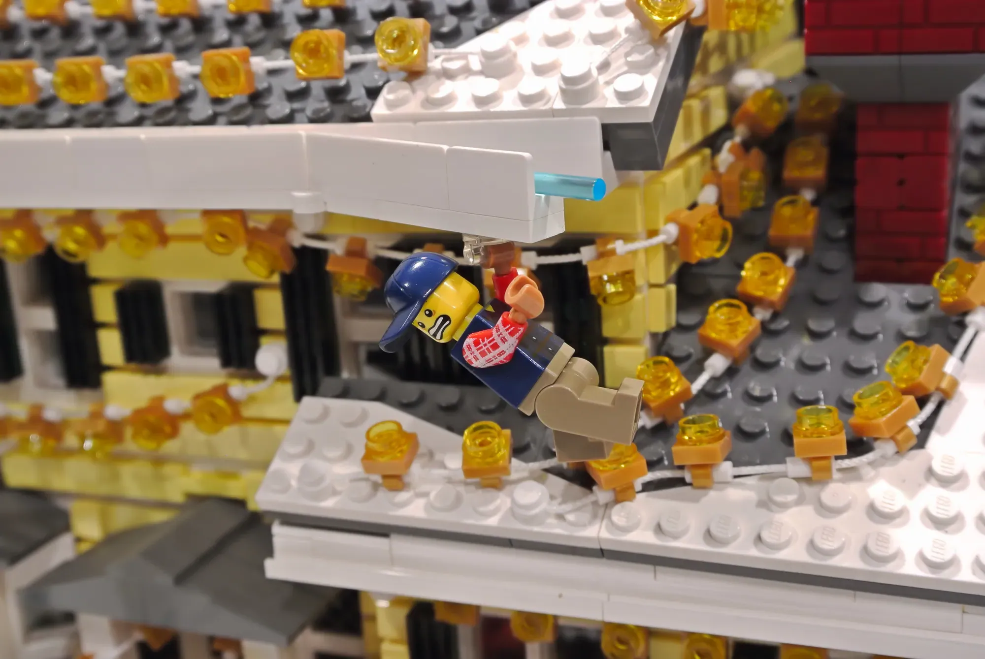 LEGO minifigure slipping on icy roof covered in golden lights during Christmas Vacation house display.