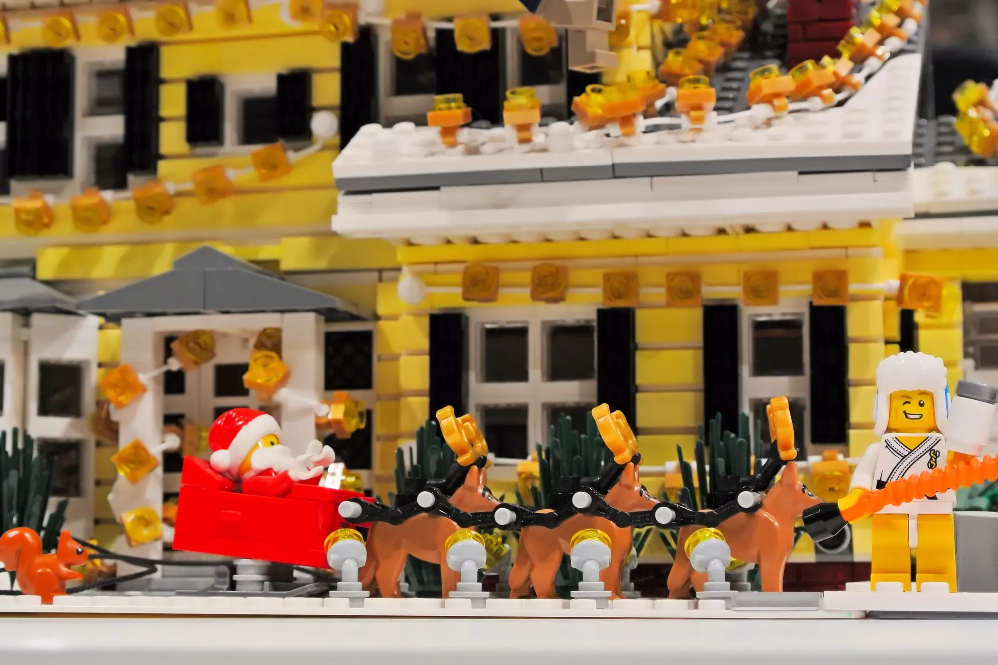 LEGO minifigure Santa with reindeer sleigh in front of elaborate yellow holiday house decorated with lights.