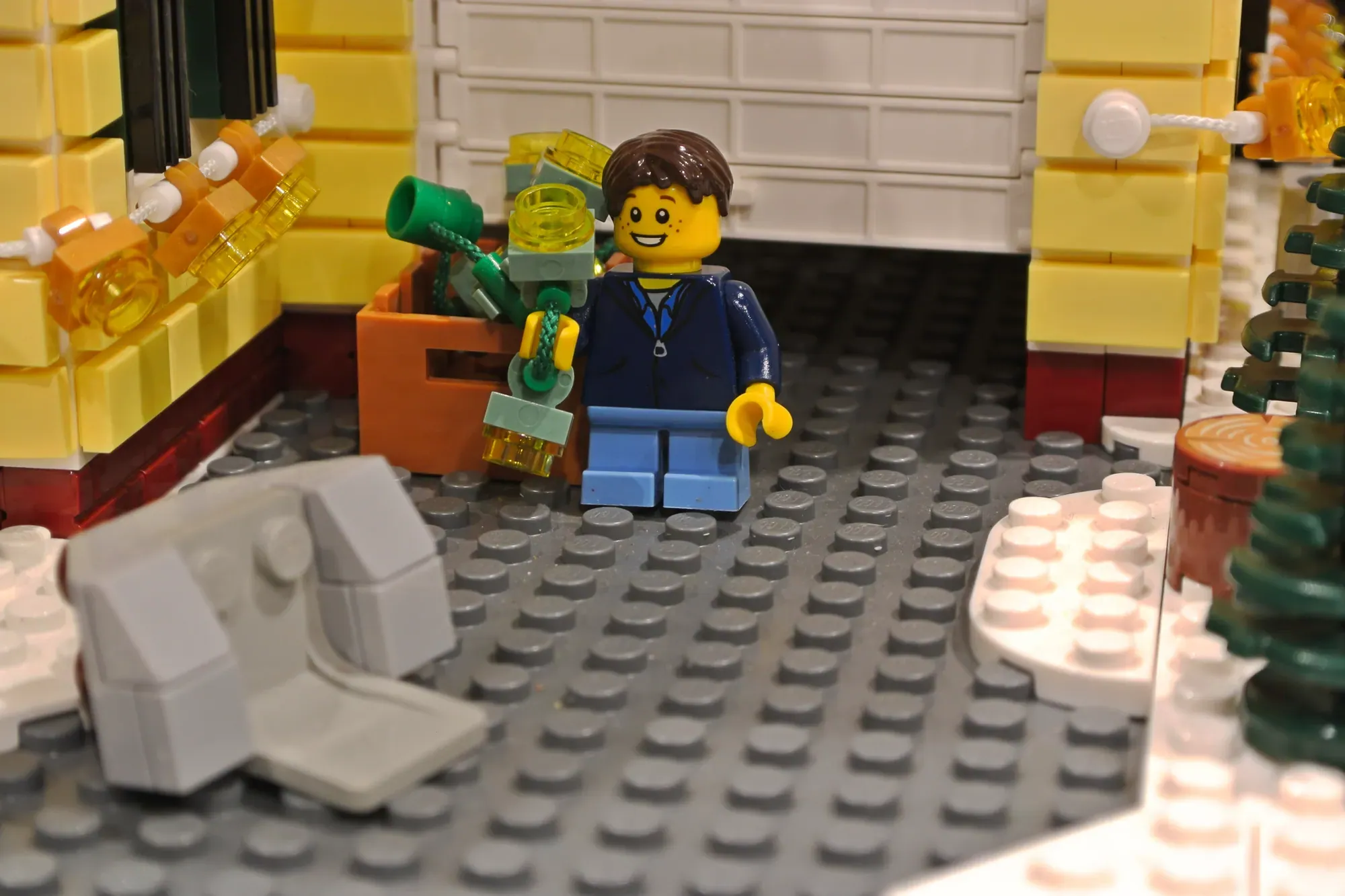 LEGO minifigure in blue jacket holding wrench inside decorated yellow house with garage and holiday lights.